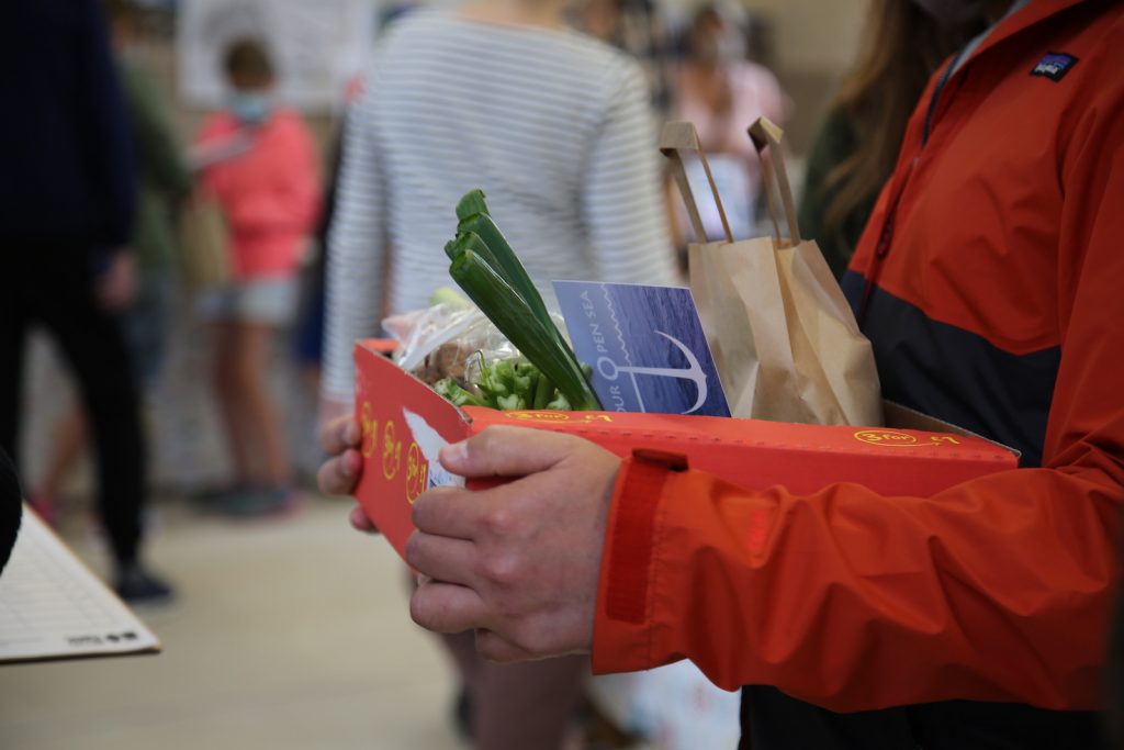 Someone holding a box of vegetables with a Safe Harbour Open Sea postcard stuck inside init