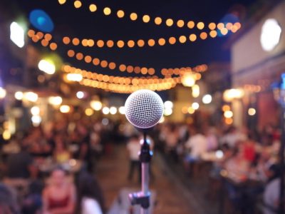 In the forefront of the picture there is a microphone awaiting a performer, in the background there are twinkly lights and an audience