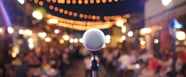 In the forefront of the picture there is a microphone awaiting a performer, in the background there are twinkly lights and an audience